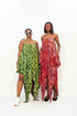 Two women modeling long, draped maxi dresses against a plain white studio background. The woman on the left wears a green dress with a white circular pattern and white sneakers.