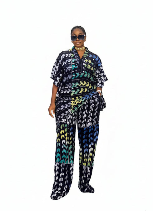 A woman modeling a matching two-piece printed outfit consisting of a short-sleeve button-up shirt and wide-leg trousers. The fabric features a bold geometric pattern in black, blue, green, yellow, and purple.
