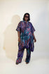 A woman modeling a matching two-piece printed outfit with a loose kaftan-style top and wide-leg pants, standing indoors against a beige wall with framed wall art and a plant. A phone screen overlay appears in the lower left showing a smaller preview of the outfit.
