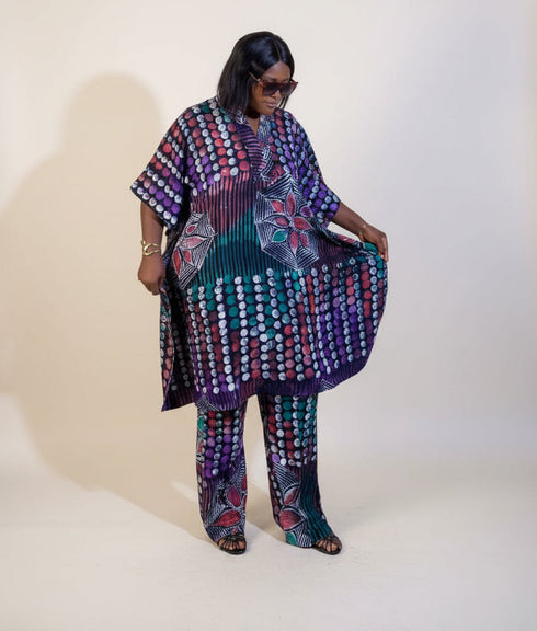 A woman models a matching two-piece printed outfit consisting of a loose short-sleeve top and wide-leg pants. The fabric features a complex pattern of purple, teal, pink, and white circular and floral motifs.