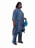 A woman modeling a blue and white patterned two-piece outfit consisting of a loose tunic top and matching pants, standing in an indoor room with a yellow wall, framed art, and a potted plant.
