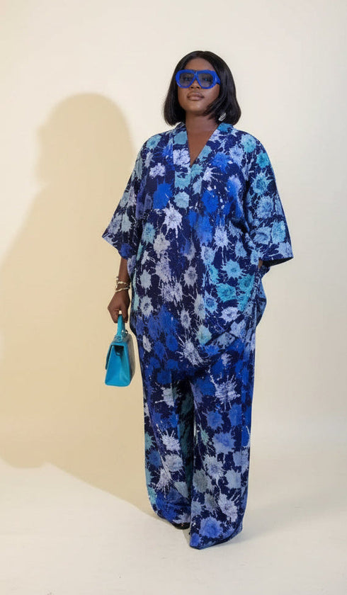 A woman wearing a blue and white floral pajama set stands in a room with framed wall art, a plant, and a green chair. A phone screen overlay in the bottom left shows a smaller version of the pajama set.