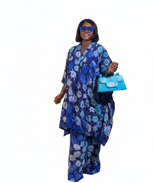 A woman wearing a blue and white floral printed kaftan tunic and matching trousers, accessorized with a bright blue handbag and blue sunglasses, posing in front of a beige wall with framed art.