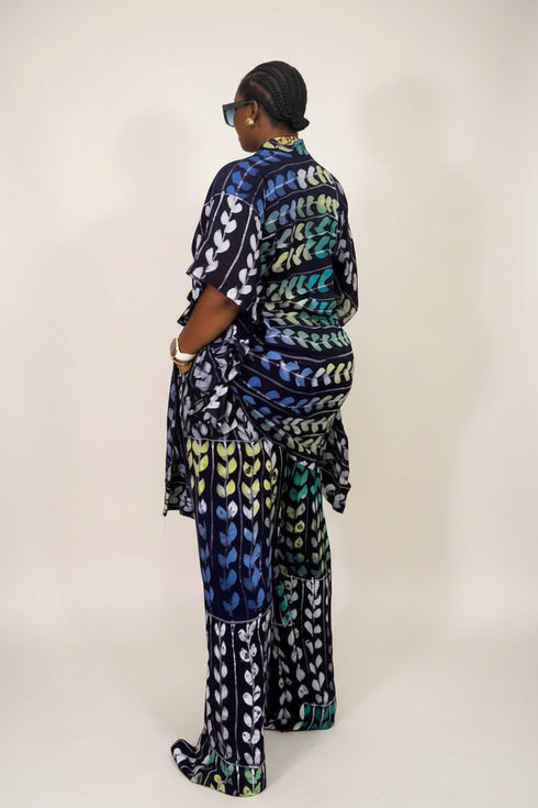 A person wearing a matching two-piece printed outfit stands in front of a striped wall. The outfit has a bold geometric pattern in blue, green, black, and white.