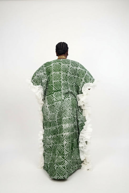A person viewed from behind wearing a green patterned kaftan dress with wide white ruffled sleeves, standing indoors in front of a black slatted wall. Large text at the top reads 'Readily available Price - N35,000 each WhatsApp +2347035008482', and a small phone screen inset appears at the bottom left.