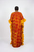 A woman viewed from behind wearing a long, flowing kaftan dress with a red and yellow floral batik pattern and dramatic ruffled yellow side panels, standing indoors against a dark striped wall.