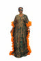 A woman modeling a long, loose-fitting patterned dress with bright orange ruffled sleeve and side details, standing in front of a black-and-white striped wall. The dress has a dark green and beige geometric print.