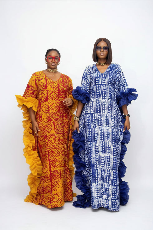 Two women modeling traditional African dress garments in a social media post. The woman on the left wears a vibrant orange and yellow patterned dress, and the woman on the right wears a blue and white patterned dress with dark blue ruffled trim.