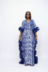 A woman modeling a long blue and white patterned kaftan dress with dramatic ruffled sleeves and hem, photographed against a plain white studio background.
