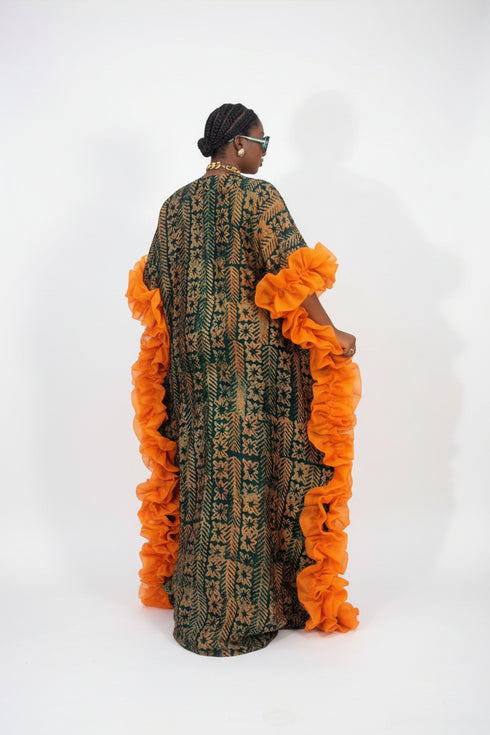 A woman wearing a long, loose caftan dress with a green and black geometric pattern and bright orange ruffled trim, shown from the back in an indoor setting with a striped wall behind her.