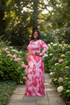 A woman stands on a stone path in a lush garden wearing a long-sleeved pink and white watercolor-patterned maxi dress with a matching belt.