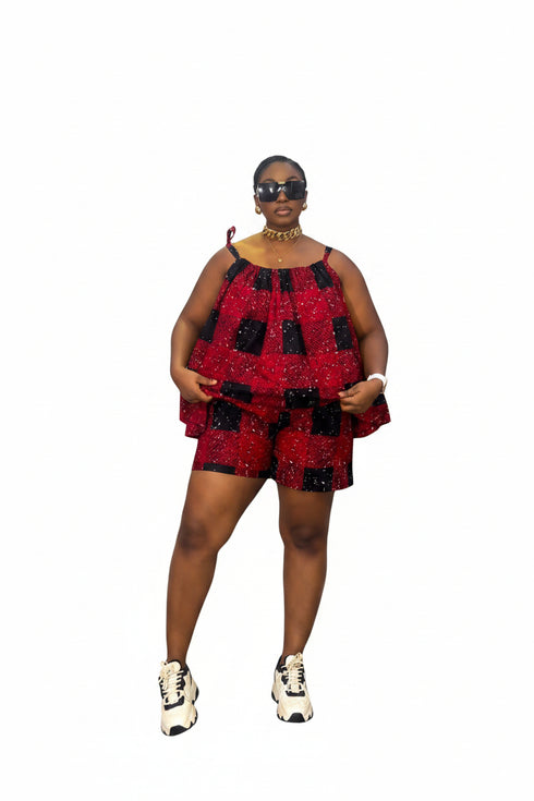 A woman modeling a red and black checkered sequined sleeveless top and matching shorts set, standing in front of a gray and white striped wall. She is wearing white and black sneakers, gold jewelry, and sunglasses.