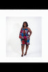 A woman modeling a colorful sleeveless two-piece matching set featuring a vibrant abstract pattern in red, blue, pink, and yellow, standing in an indoor setting with a beige wall, framed prints, and a potted plant.