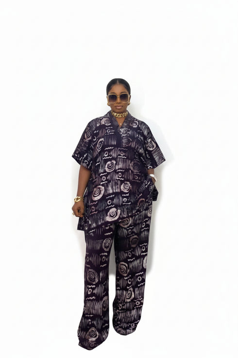A person wearing a matching two-piece outfit with a dark purple and black abstract floral pattern, standing in front of a black and white striped wall.