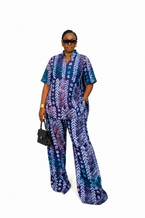 A woman modeling a matching short-sleeve button-front top and wide-leg trousers set in a blue, purple, and teal geometric pattern, styled with sunglasses, a gold choker, a gold bracelet, and a small black handbag against a plain white background.