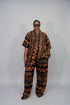 A person modeling a matching short-sleeve button-up shirt and wide-leg pants set with a bold circular and geometric pattern in orange, brown, and black, photographed against a plain light gray studio background.