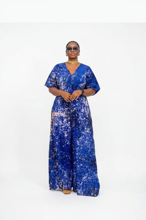 A woman modeling a royal blue and white patterned short-sleeve jumpsuit in front of a gray slatted wall. The image is a screenshot from a social media post with text overlays showing a price and WhatsApp contact.
