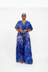 A woman modeling a royal blue and white patterned short-sleeve jumpsuit in front of a gray slatted wall. The image is a screenshot from a social media post with text overlays showing a price and WhatsApp contact.