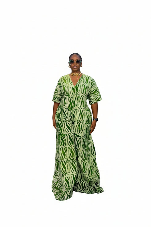 A woman modeling a green and black patterned short-sleeve jumpsuit in front of a black slatted wall. The image is a screenshot from a social media post with text overlays showing a price and WhatsApp contact.