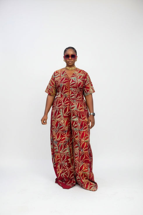 A woman modeling a red and orange patterned short-sleeve jumpsuit in an indoor setting with a striped wall background. Text overlays indicate the price is N32,000 each and provide a WhatsApp contact.