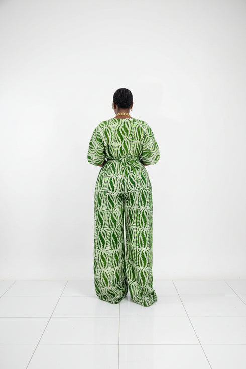 A person shown from the back wearing a green and white patterned short-sleeve jumpsuit in front of a black slatted wall. Text overlays show pricing and WhatsApp contact information.