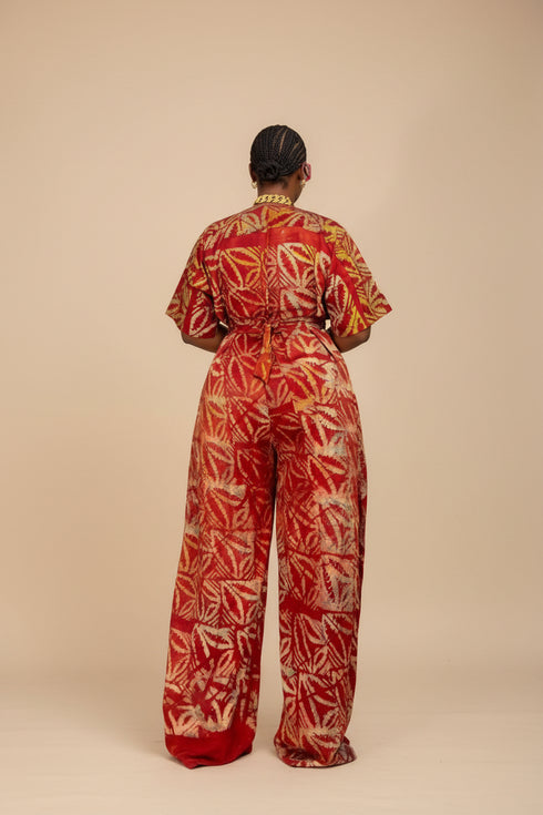 A woman wearing a red and orange patterned traditional dress stands in front of a black slatted wall, shown within a social media post layout with text overlays advertising availability and price.