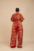 A woman wearing a red and orange patterned traditional dress stands in front of a black slatted wall, shown within a social media post layout with text overlays advertising availability and price.