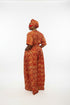 A woman wearing a red and orange patterned traditional outfit stands in front of a gray slatted wall, with overlaid text showing a price and WhatsApp contact. This is a social-media-style listing image rather than a clean product photo.