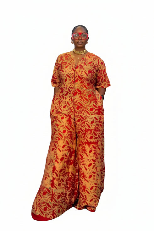 A woman modeling a long-sleeved, wide-leg jumpsuit with a bold red and gold abstract pattern, standing in front of a black and white striped wall. A phone screen overlay is visible in the bottom-left corner showing a video preview of the garment.