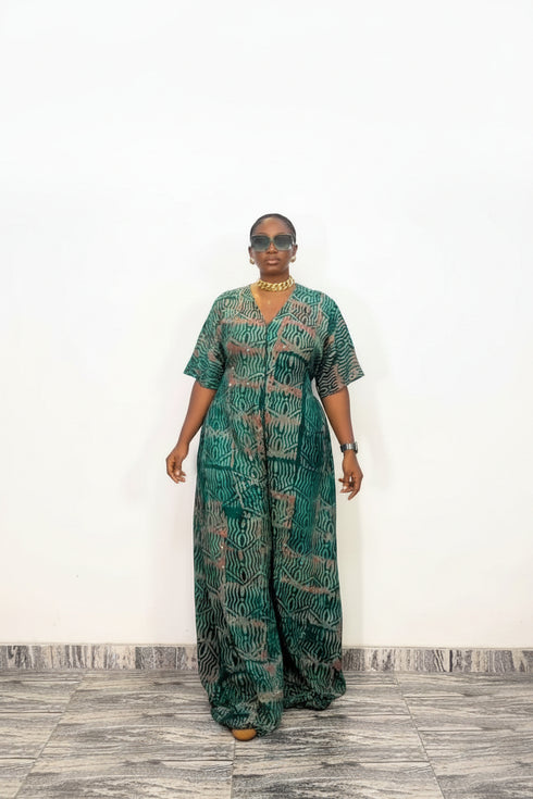 A woman modeling a green and black patterned kaftan dress with short sleeves and a V-neckline, standing against a plain white wall.
