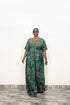 A woman modeling a green and black patterned kaftan dress with short sleeves and a V-neckline, standing against a plain white wall.