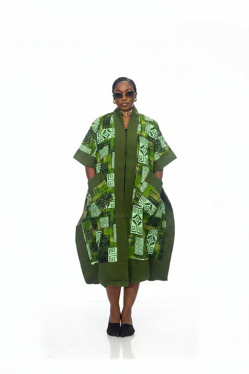 A woman modeling a green patterned kaftan dress with short sleeves, pockets, and a high collar, standing against a plain white background.