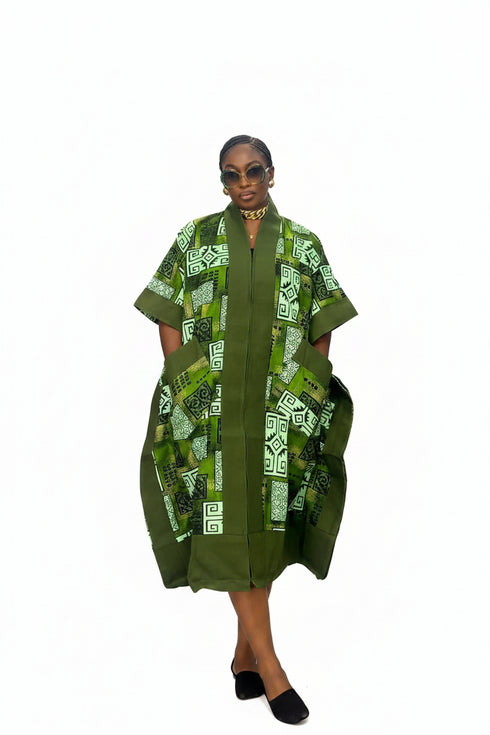 A woman modeling a green and white patterned kaftan dress with a high collar, short sleeves, and front pockets, set against a plain white background.