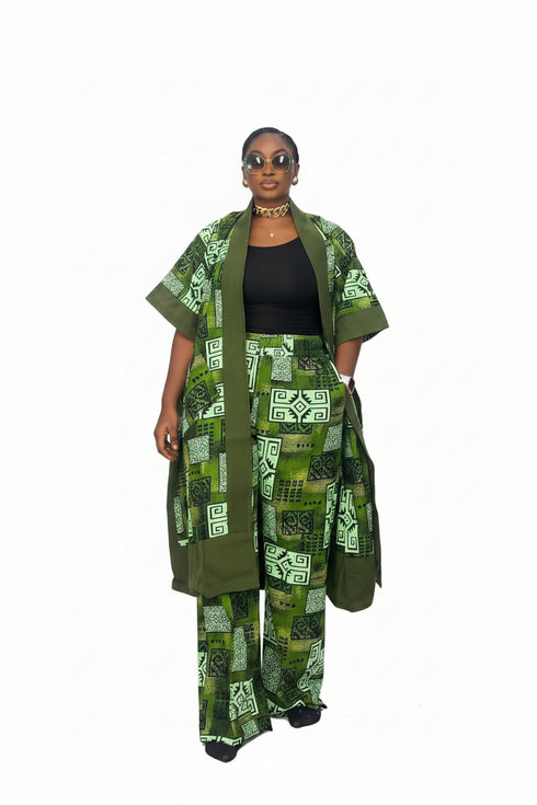 A woman modeling a green and black patterned African wax print outfit, consisting of a matching jacket and trousers with a black top underneath, set against a black and white striped background.